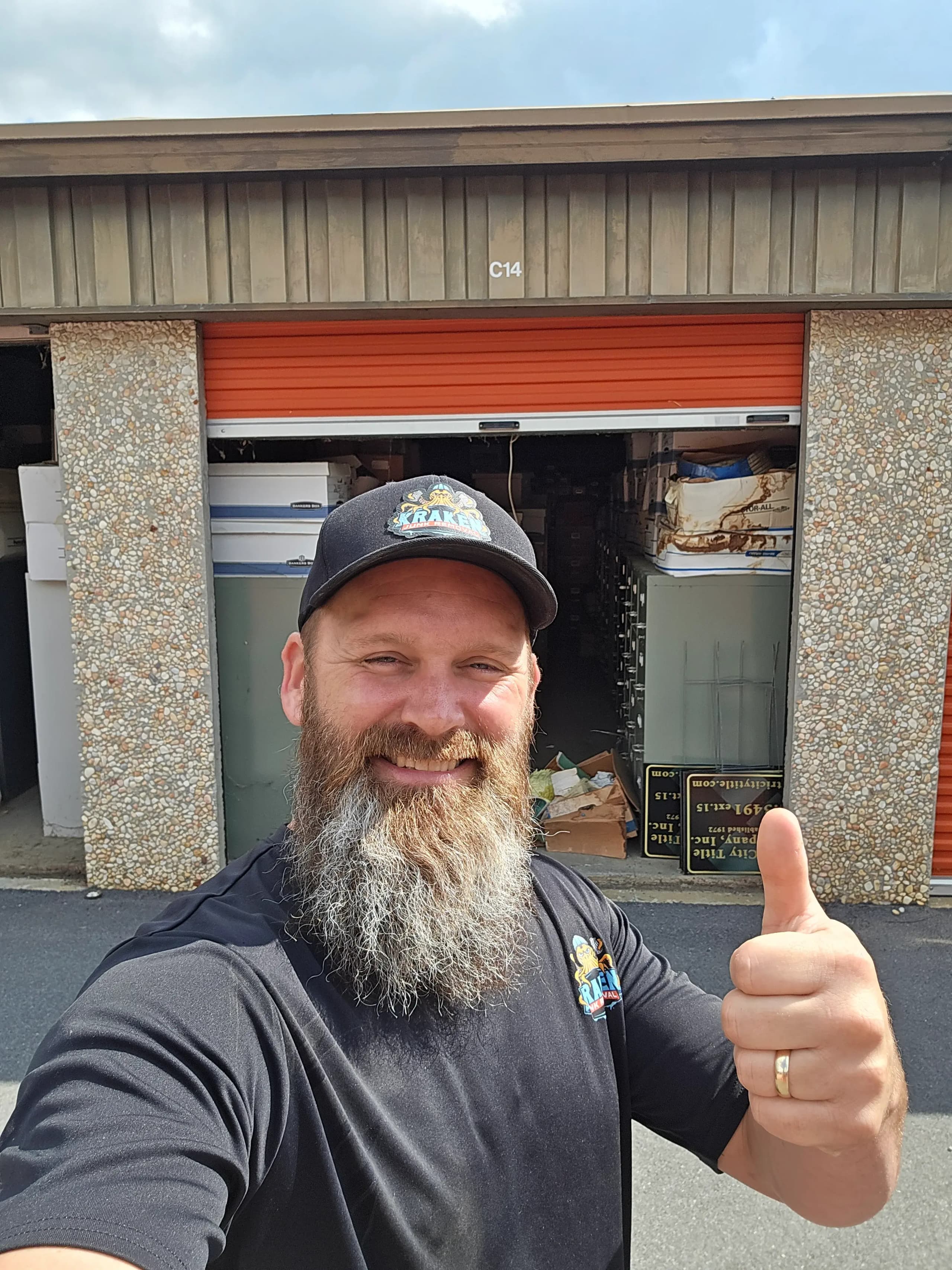 Kraken Junk Removal crew member giving a thumbs-up in front of an open storage unit packed with file boxes and metal cabinets