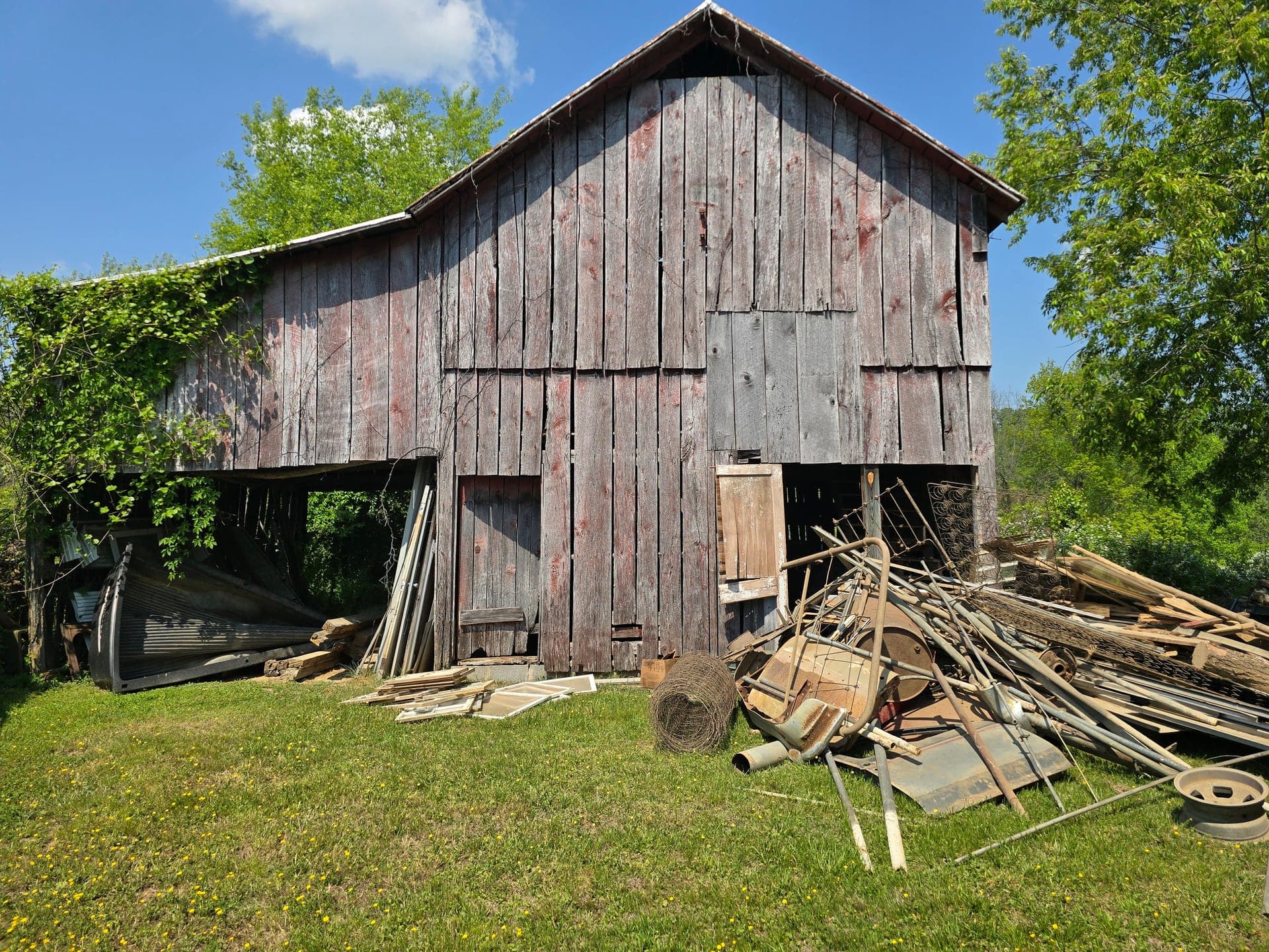 Weathered wooden barn with scrap metal, lumber, and mixed debris piled in the yard and open bays before removal