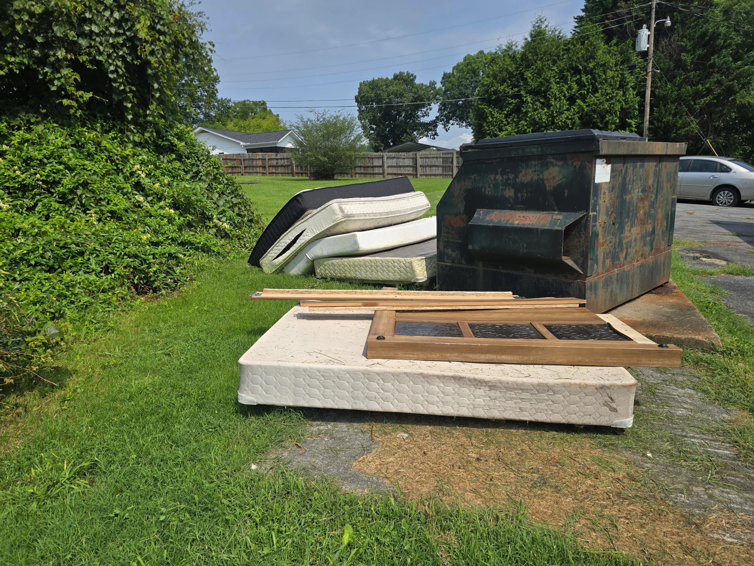 Discarded mattresses and bed-frame pieces beside a dumpster before cleanup.