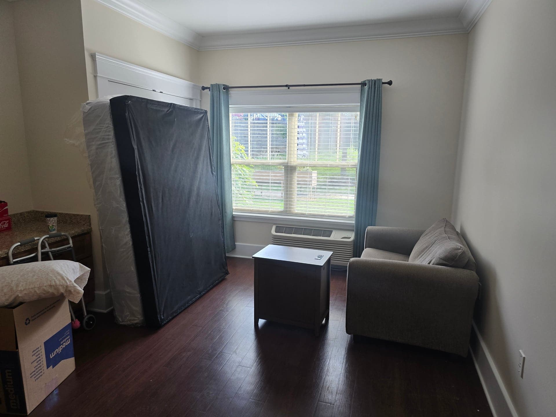 Plastic-wrapped mattress, gray sofa, side table, and cardboard box staged in a room during an estate cleanout