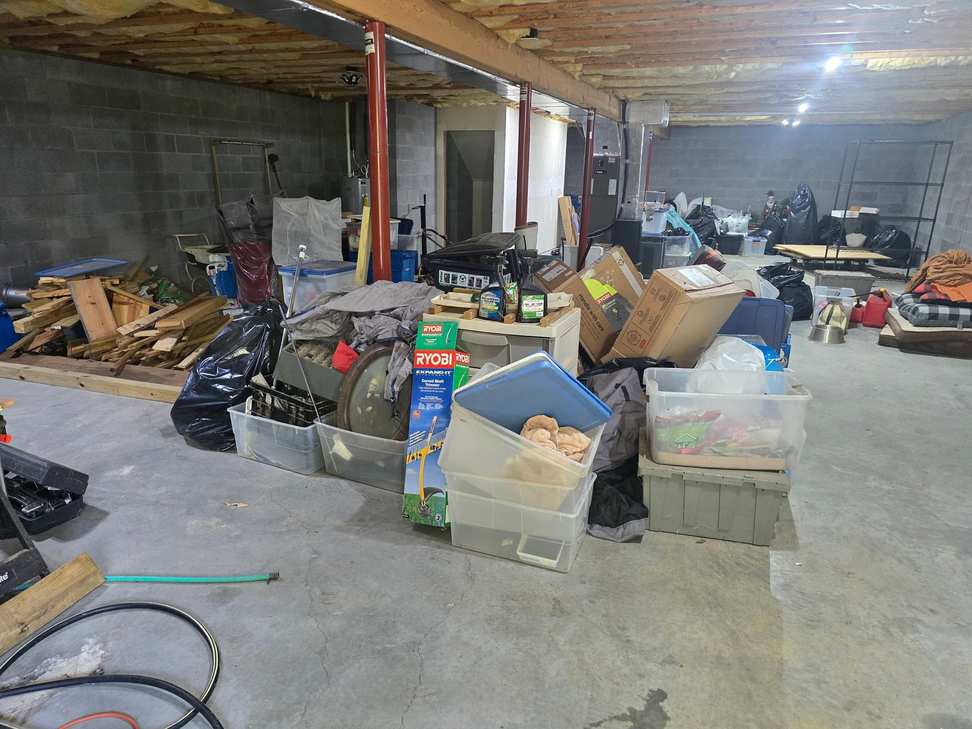 Cluttered unfinished basement filled with storage bins, boxes, and household items during an estate cleanout