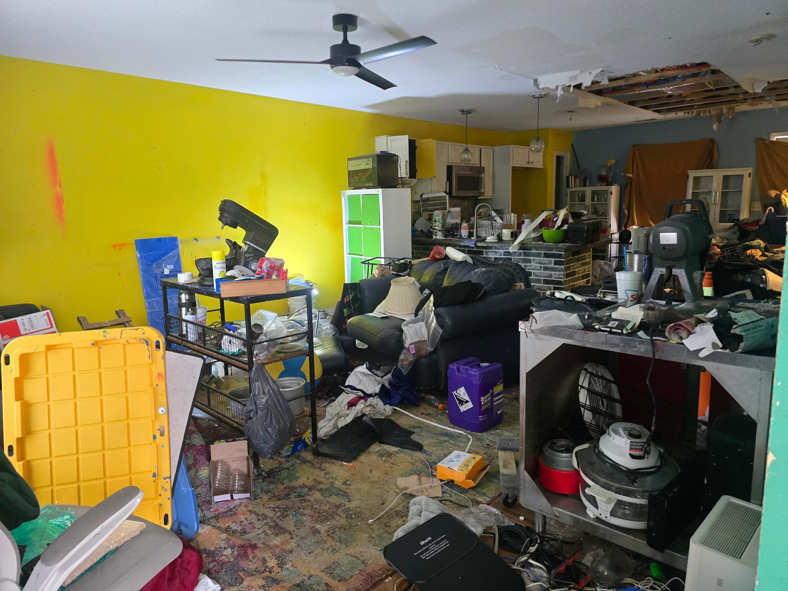 Living room and kitchen filled with a couch, shelves, appliances, and household clutter, with a hole in the ceiling