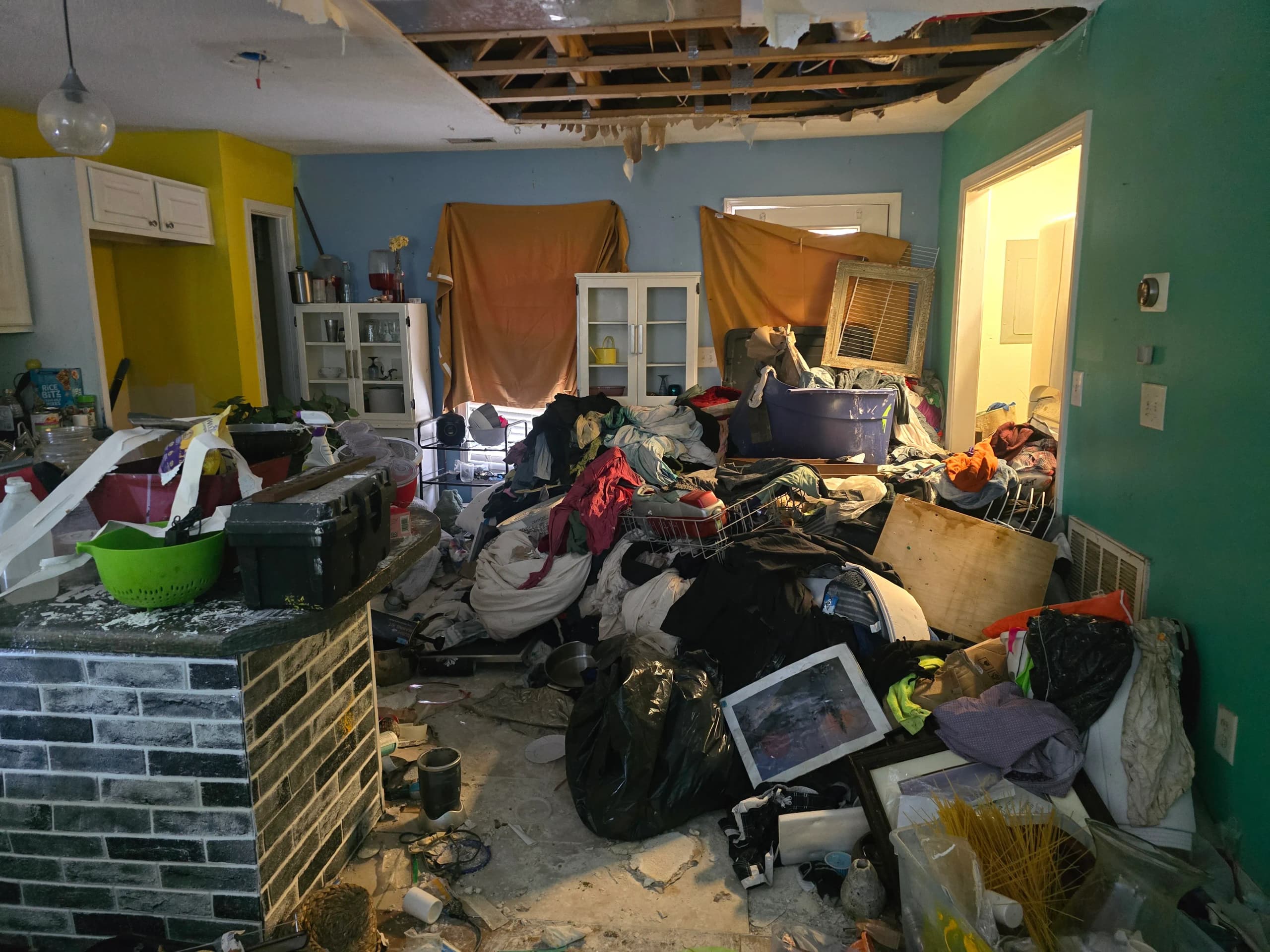 Kitchen with a brick island, clothes and bags piled across the floor, and an open hole in the ceiling