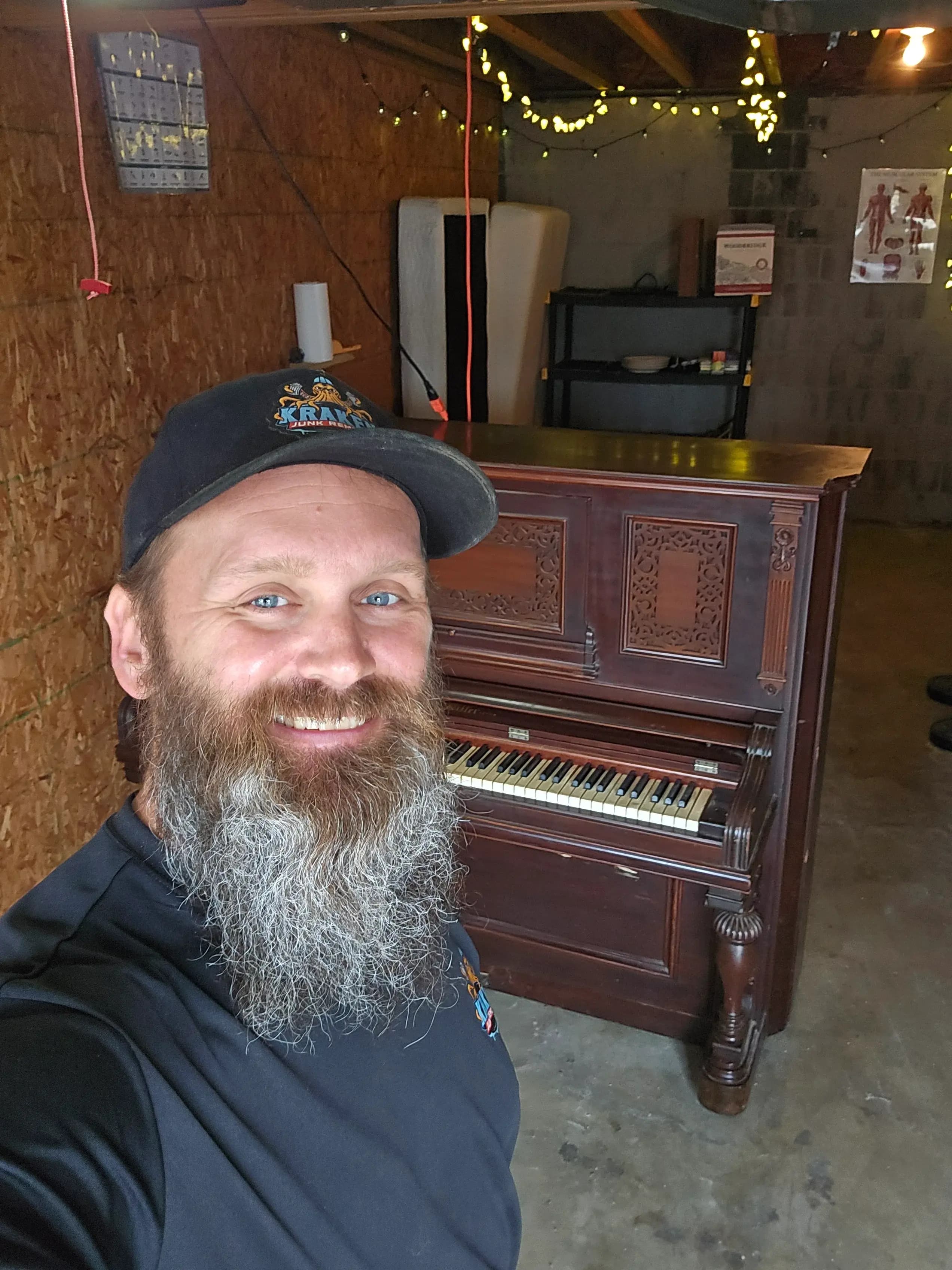Kraken Junk Removal crew member smiling beside a dark wood upright piano ready for removal
