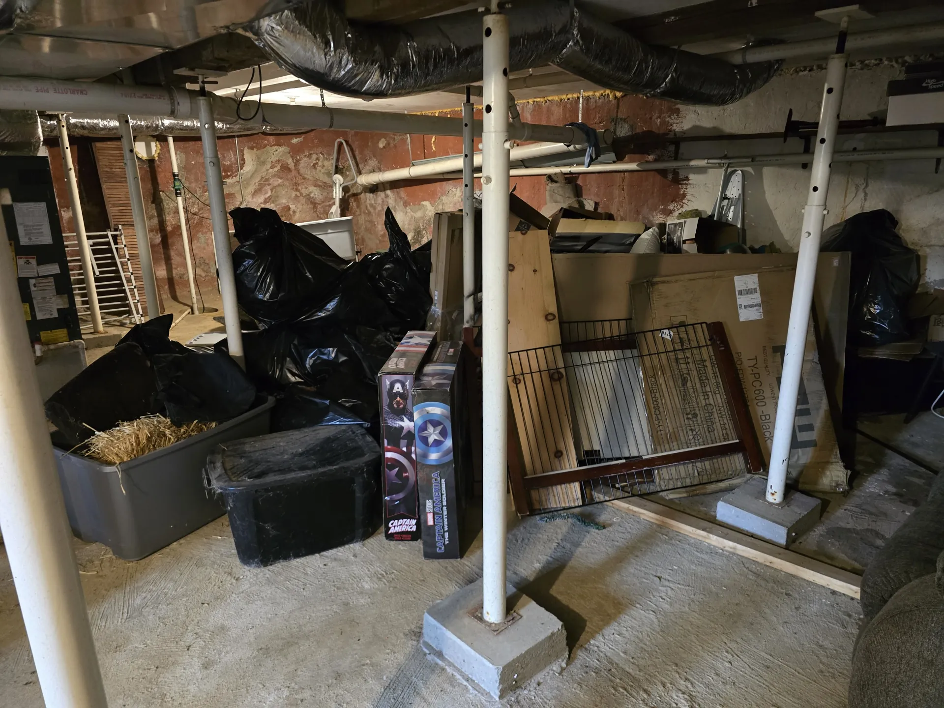 Unfinished basement with low joists, support posts, trash bags, boxes, and household clutter